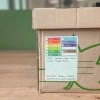 Une boîte en carton avec une étiquette indiquant son contenu et l'adresse de livraison. Les catégories sur l'étiquette incluent cuisine, salon, salle à manger, garage, buanderie, jardin, et d'autres, avec des éléments comme des classeurs et des stylos listés comme contenu.