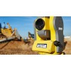 L'image montre un appareil de mesure de terrain de couleur jaune sur un chantier, avec des équipements de construction flous en arrière-plan.