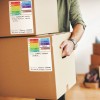L'image montre une personne portant des cartons étiquetés avec différents contenus. Les étiquettes indiquent des catégories comme 'cuisine', 'salon', et 'chambre'. Le carton contient des 'livres, doudous, jouets...'.