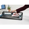 L'image montre une personne utilisant un outil de découpe sur un bureau, avec un écran d'ordinateur et une tasse à proximité.