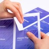 L'image montre une personne en train de décoller une étiquette ou un autocollant d'une feuille, avec des noms sur des zones colorées en violet au fond.