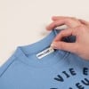 L'image montre un pull bleu avec une étiquette portant le nom 'Marin's Guyot' et un motif de texte noir qui dit 'VIE E'. Une main tient le col du pull.