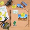L'image montre un carnet de souvenirs de voyage avec des photos, un appareil photo, des ciseaux et des étiquettes. Le carnet est décoré de dessins de cactus et de mots en français.