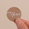 L'image montre un petit cercle en papier avec le mot 'Merci' écrit en blanc sur un fond brun. La main qui tient le cercle est visible.