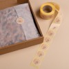 Cette image montre une boîte en carton avec du papier de soie à l'intérieur, étiquetée avec un autocollant rond. À côté, un rouleau d'étiquettes autocollantes est visible.